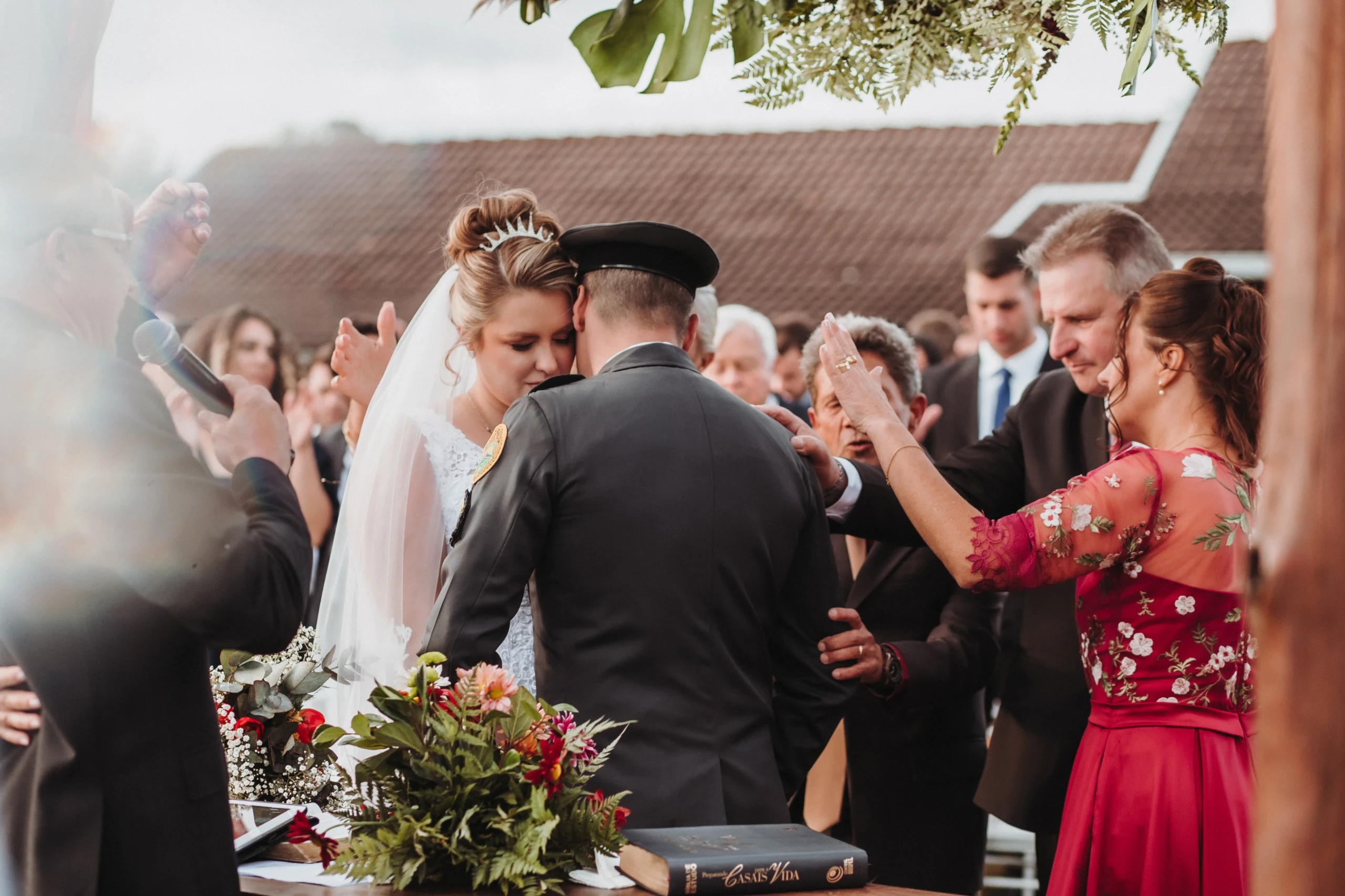 Bruidspaar waarvan de man gekleed in uniform omhelst elkaar, omring door familie.