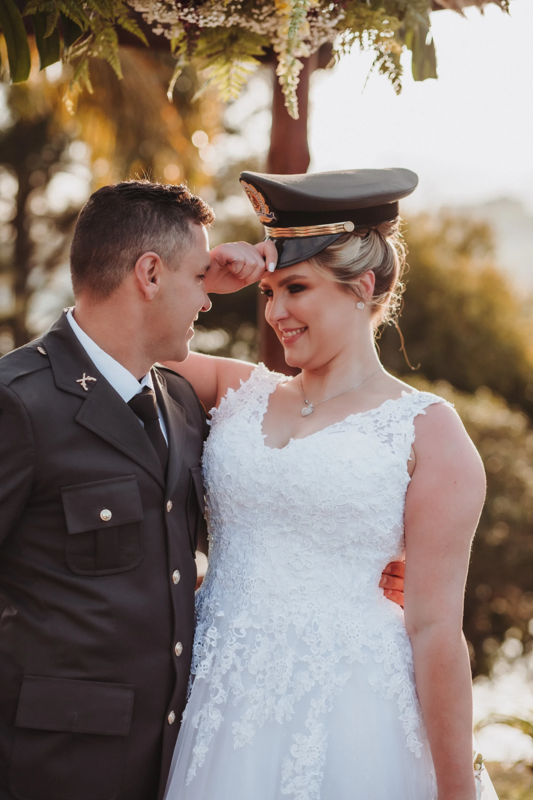 Vrolijk bruidspaar waarvan de man gekleed in uniform. De bruid draagt een wit kanten jurk met een bruidsboeket bestaande uit witte, rode en gele bloemen. De bruid draagt ook de hoed van het uniform van de bruidegom.