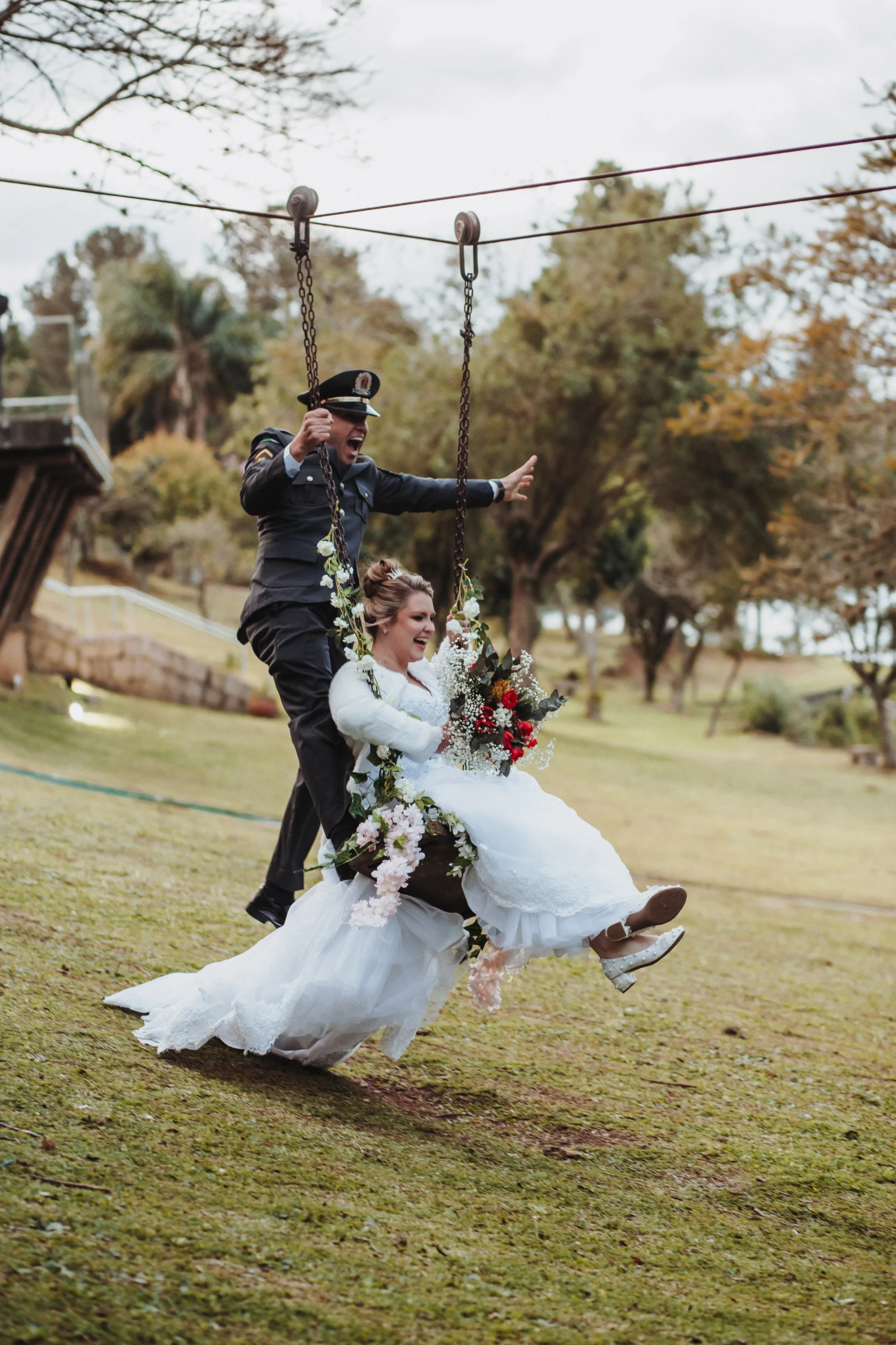 Vrolijk bruidspaar waarvan de man gekleed in uniform. De bruid draagt een wit kanten jurk met een bruidsboeket bestaande uit witte, rode en gele bloemen. De bruid zit op een schommel en de bruidegom staat met 1 voet op de schommel terwijl hij zich vasthoudt aan de ketting.