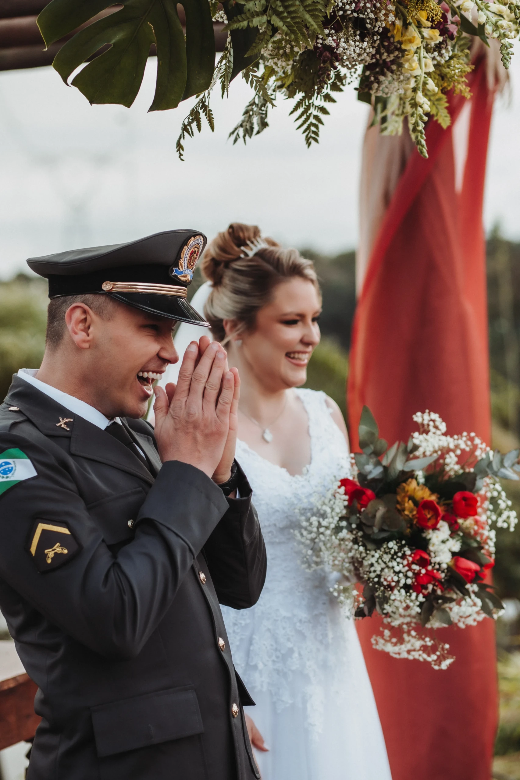 Vrolijk bruidspaar waarvan de man gekleed in uniform. De bruid draagt een wit kanten jurk met een bruidsboeket bestaande uit witte, rode en gele bloemen.