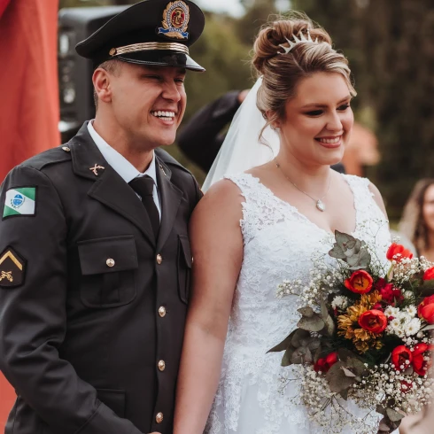 Vrolijk bruidspaar waarvan de man gekleed in uniform. De bruid draagt een wit kanten jurk met een bruidsboeket bestaande uit witte, rode en gele bloemen.