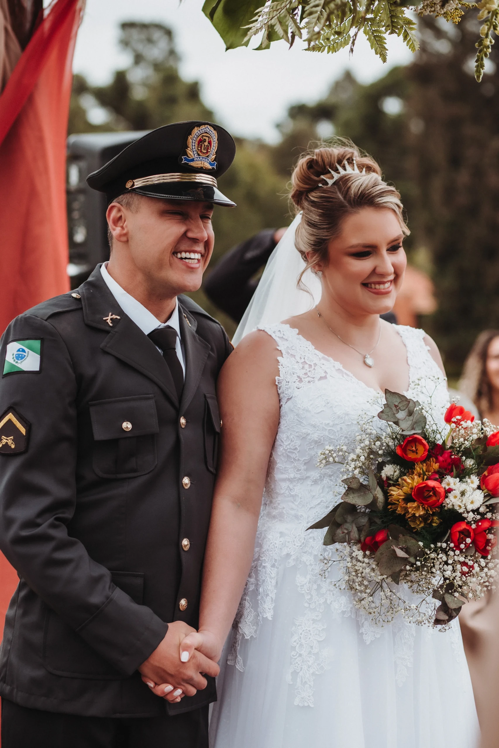Vrolijk bruidspaar waarvan de man gekleed in uniform. De bruid draagt een wit kanten jurk met een bruidsboeket bestaande uit witte, rode en gele bloemen.