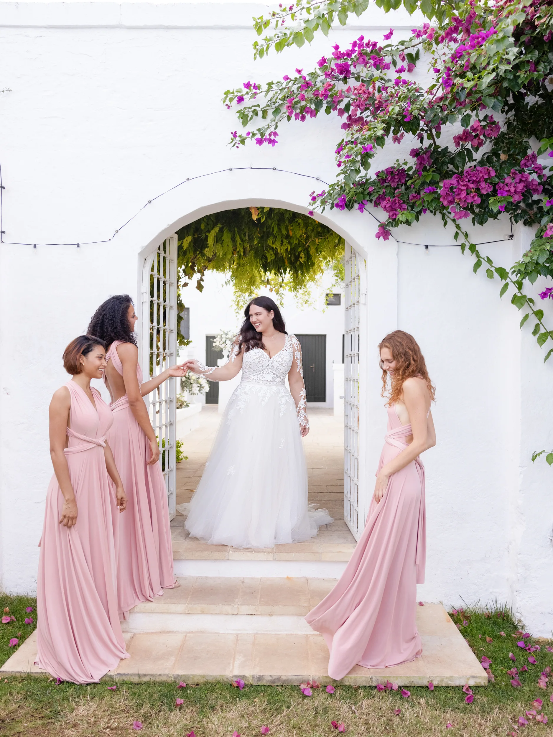 Een plussize bruid stapt een tuin in waar drie brides of honour in een roze jruk staan. Bruid draagt een witte trouwjurk van Modeca uit de collectie Victoria Collection_Veronica