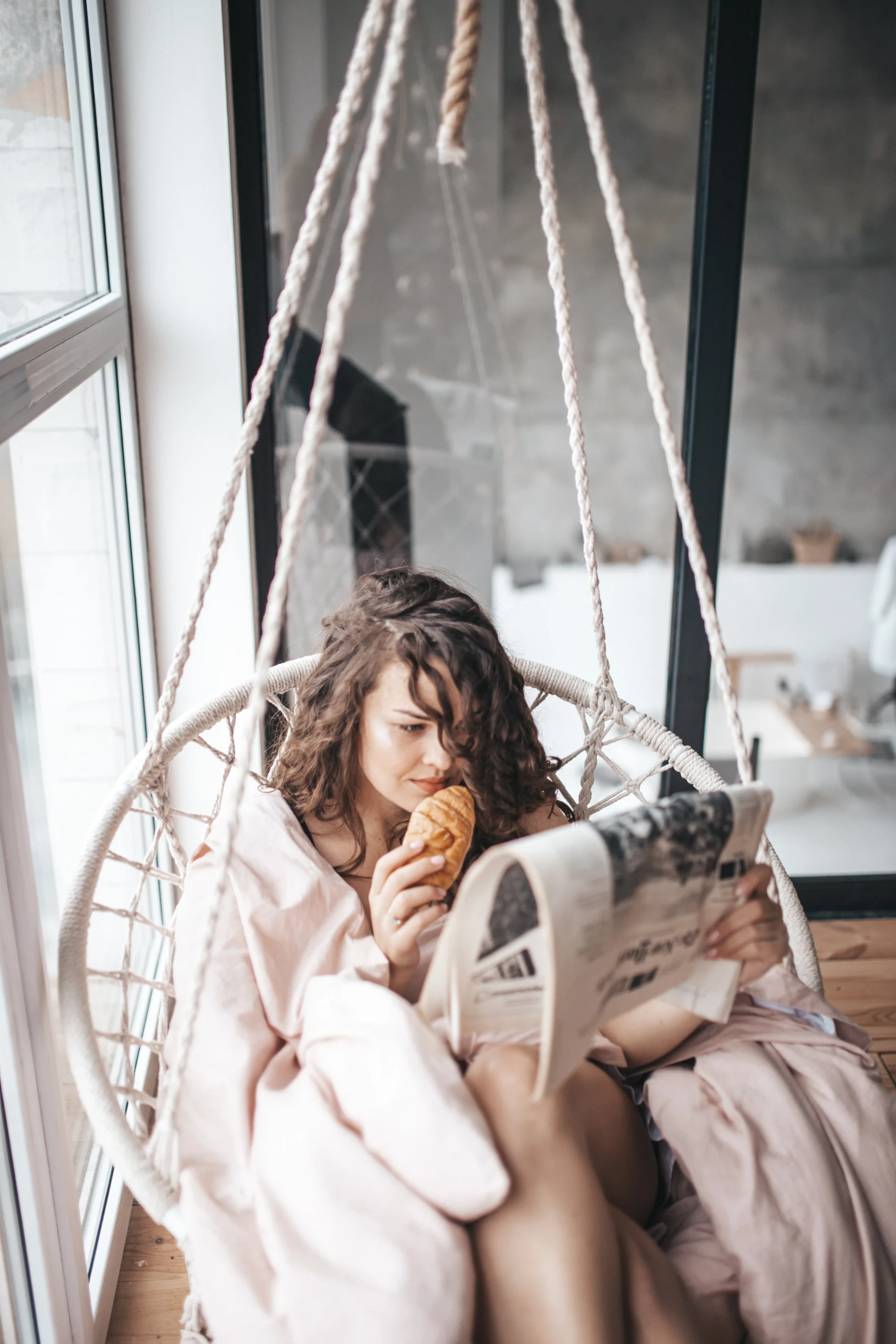 Vrouw ontspant in een hangstoel met enen krant en croissant.