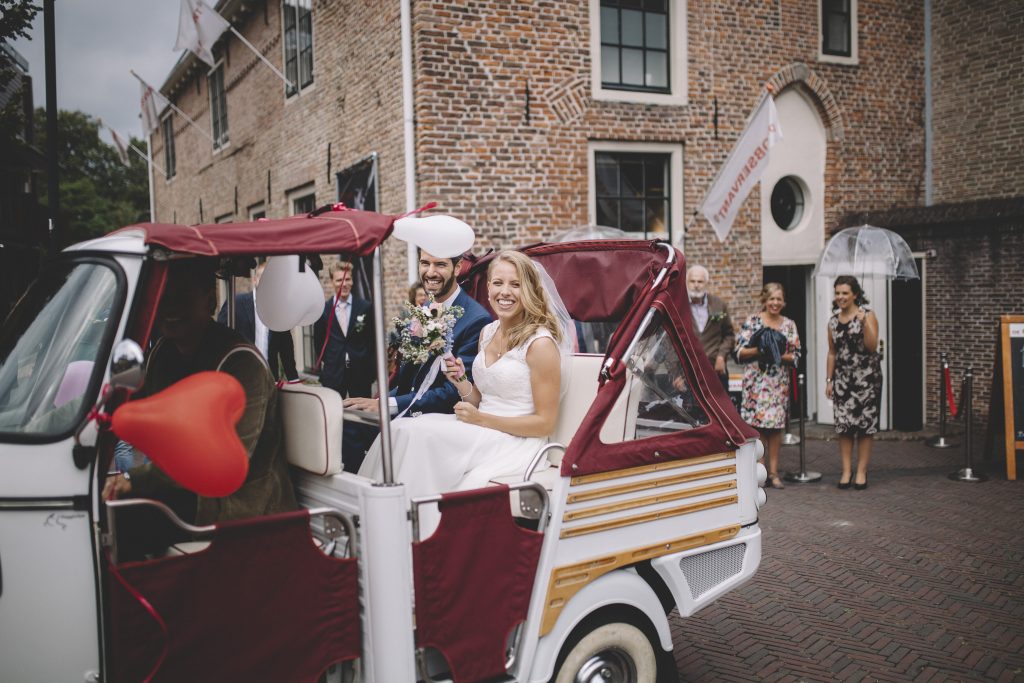 Bruid en bruidegom lachen terwijl ze in een versierde tuktuk zitten, omringd door gasten voor een historisch gebouw.