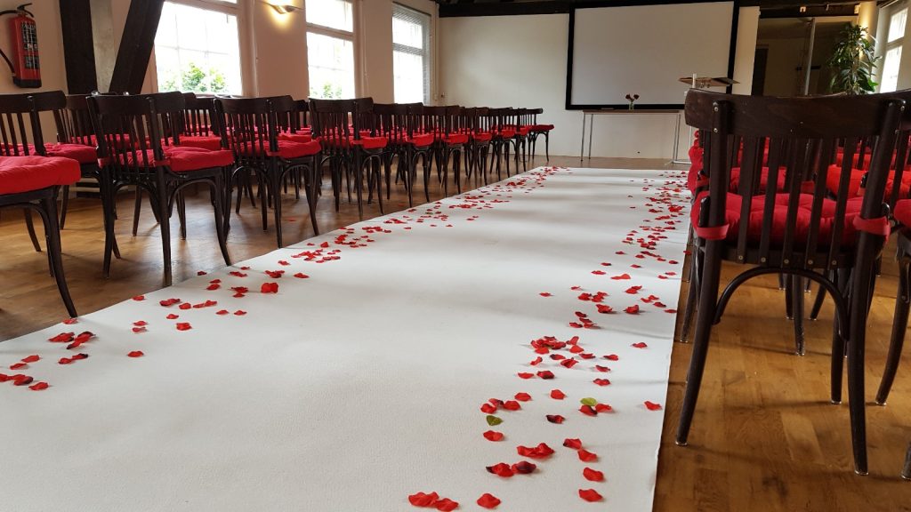 Versierde trouwlocatie met een wit gangpad bedekt met rode rozenblaadjes, omringd door houten stoelen met rode kussens.