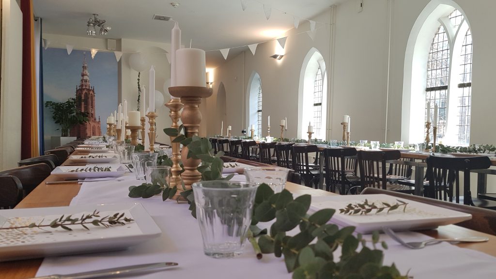 Stijlvol gedekte dinertafels met witte lopers, gouden kandelaars en groene eucalyptusdecoratie in een sfeervolle zaal met hoge ramen.
