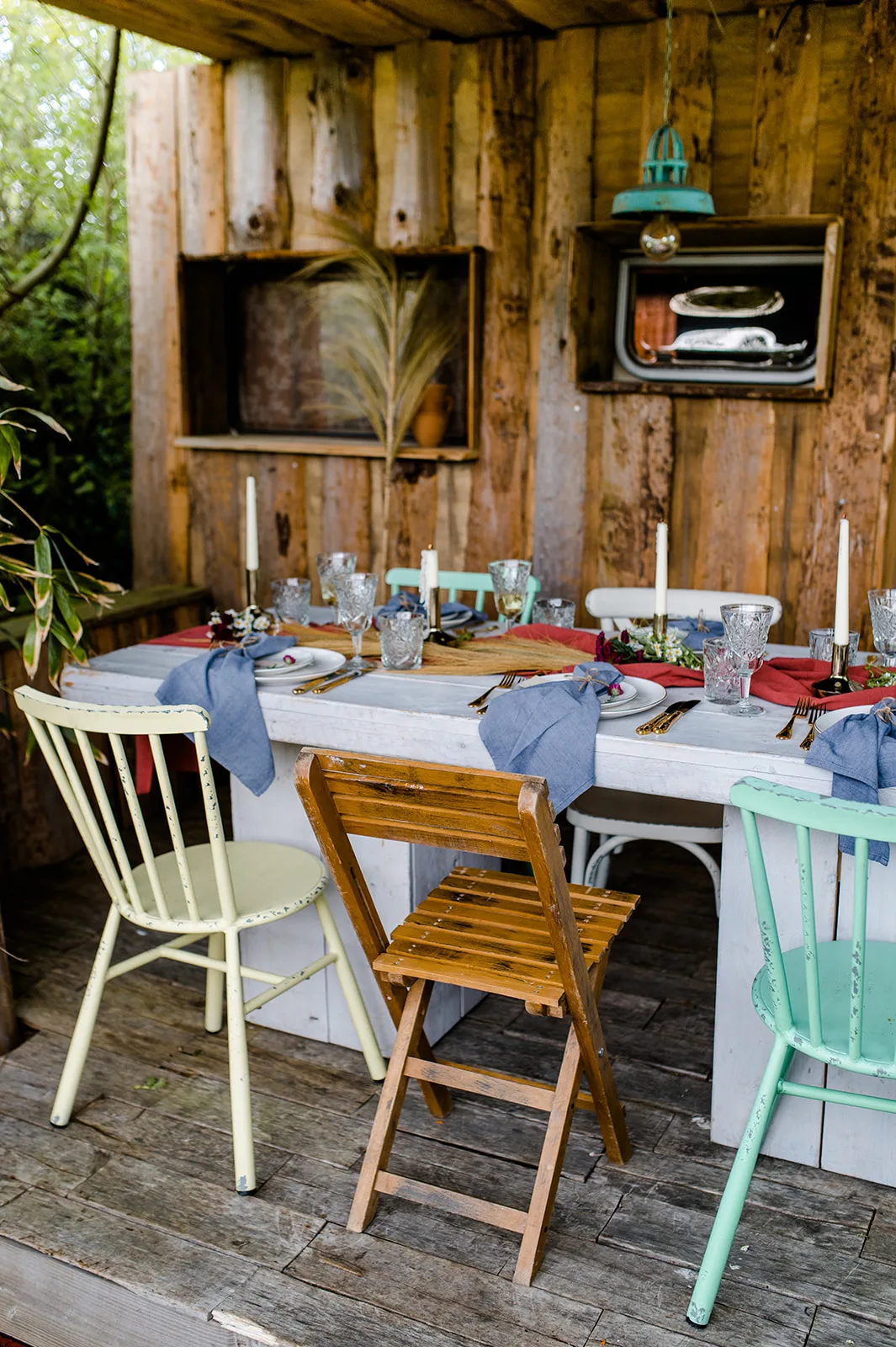 Houten hut buiten met eettafel en verschillende vintage stoelen.