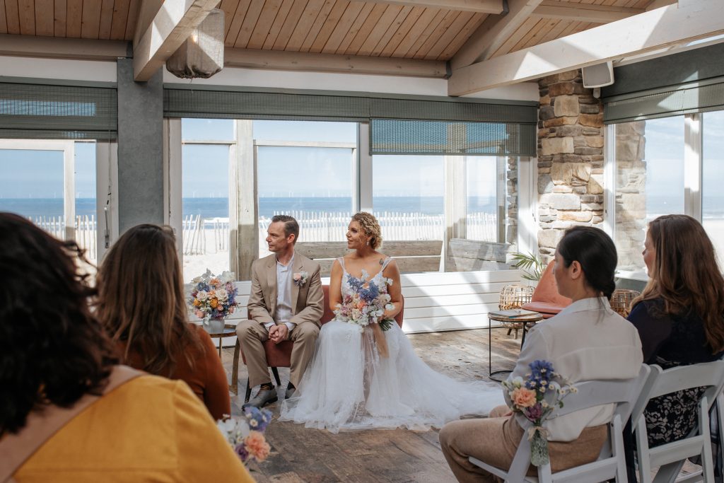 Een bruid en bruidegom zitten op een bankje tijdens hun ceremonie. De ceremonie is binnen bij een strandpaviljoen met de zee op de achtergrond.
