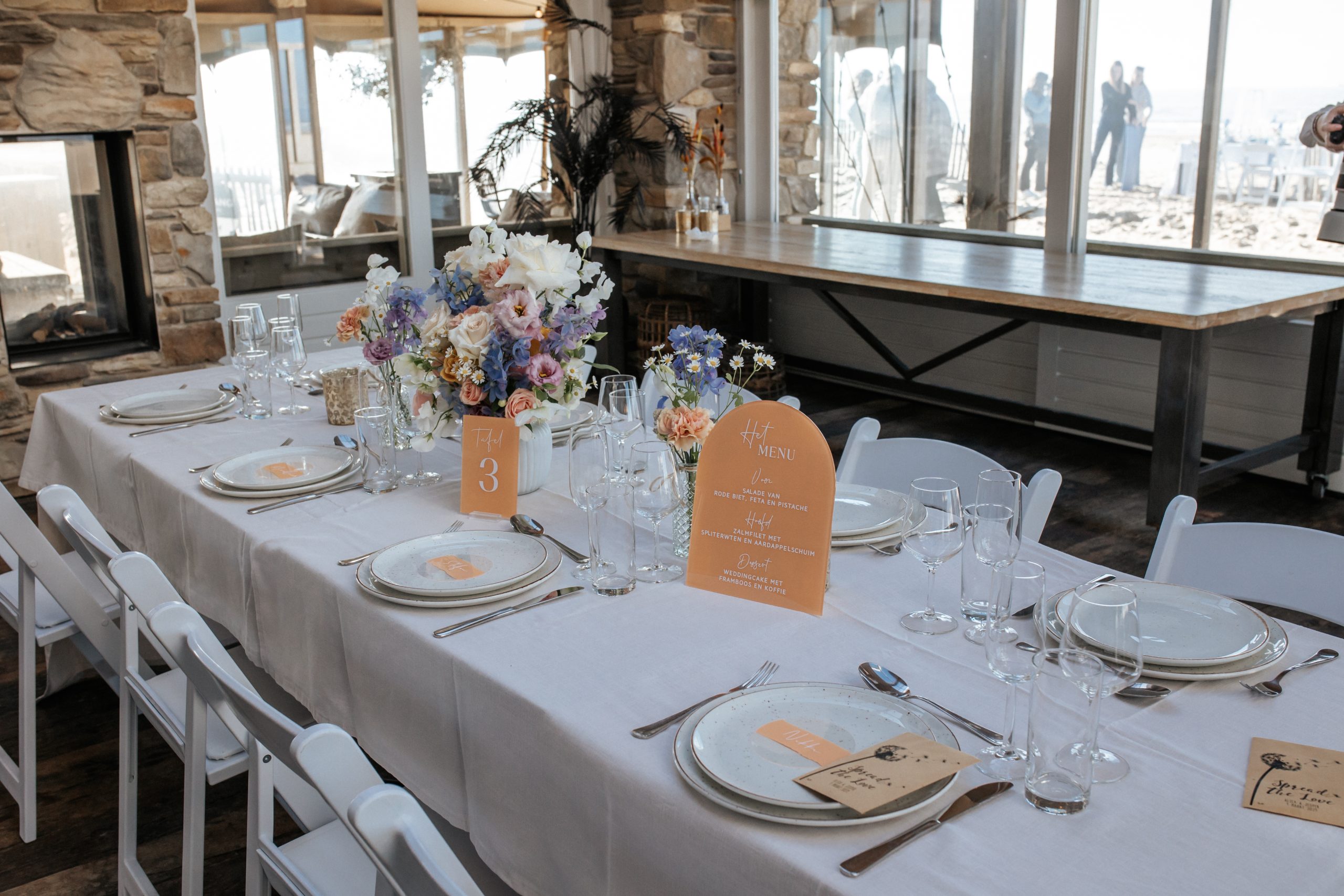 Een stijlvol gedekte tafel met witte tafellakens, servies, glazen en bestek in een lichte ruimte. Op de tafel staan kleurrijke bloemen in zachte pasteltinten, een menukaart en een tafelnummertje met "Tafel 3". In de achtergrond is een open haard en uitzicht op het strand te zien.