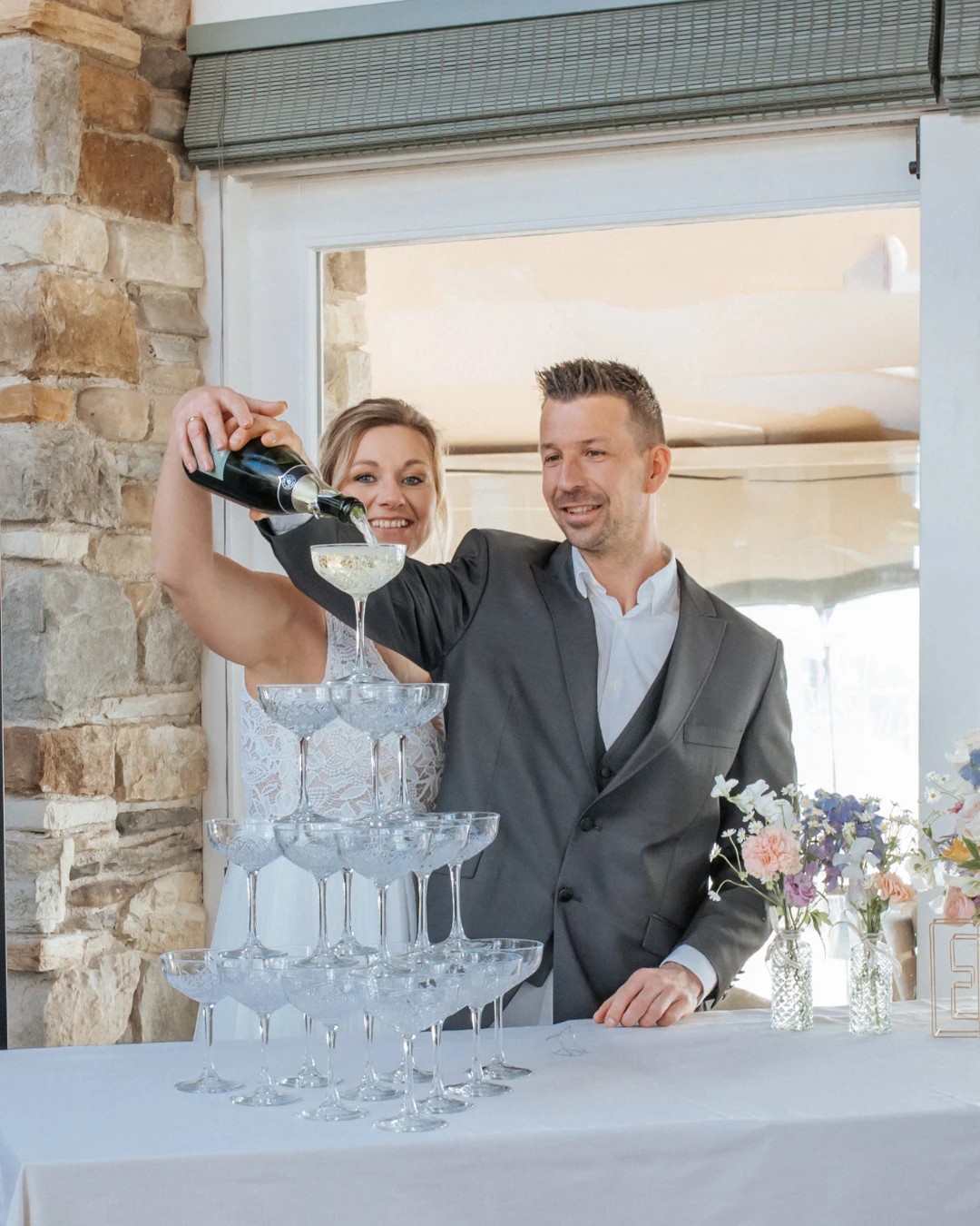 Een bruid en bruidegom schenken samen champagne in een toren van glazen tijdens hun bruiloft. Op de achtergrond is een stenen muur en een decoratief bord met de tekst "This way to the Champagne" te zien.