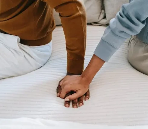 Twee mensen die met de rug naar elkaar toe op bed zitten en elkaar hand vasthouden.