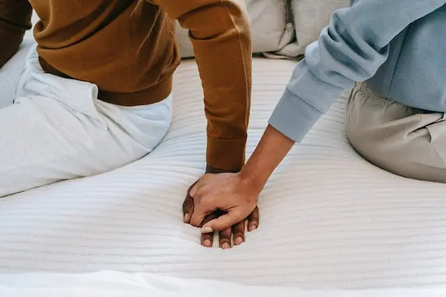 Twee mensen die met de rug naar elkaar toe op bed zitten en elkaar hand vasthouden.