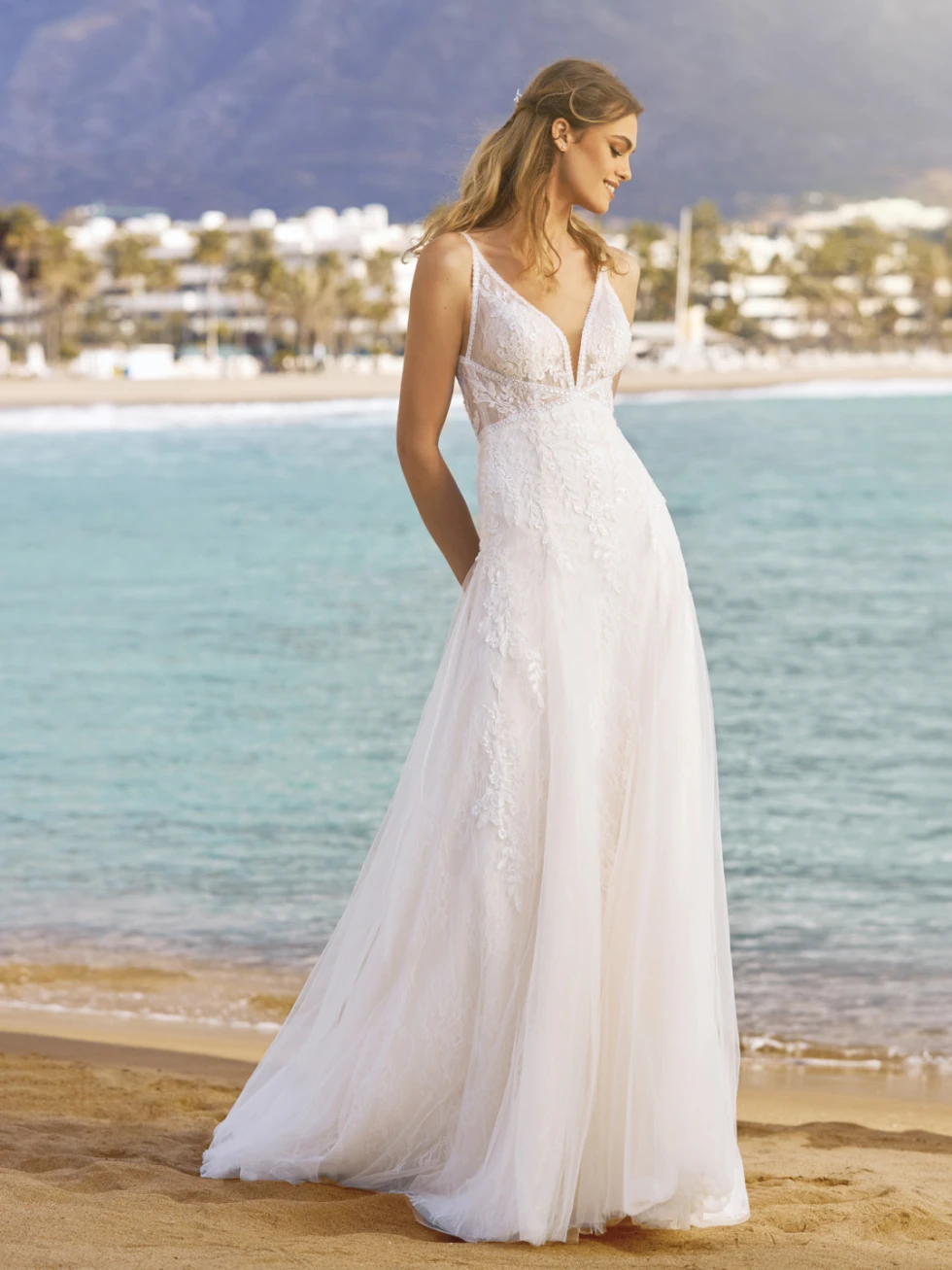 Model met A-lijn trouwjurk aan. Poseert op het strand alsof ze loopt. De jurk heeft een sweetheart halslijn en chantilly kant