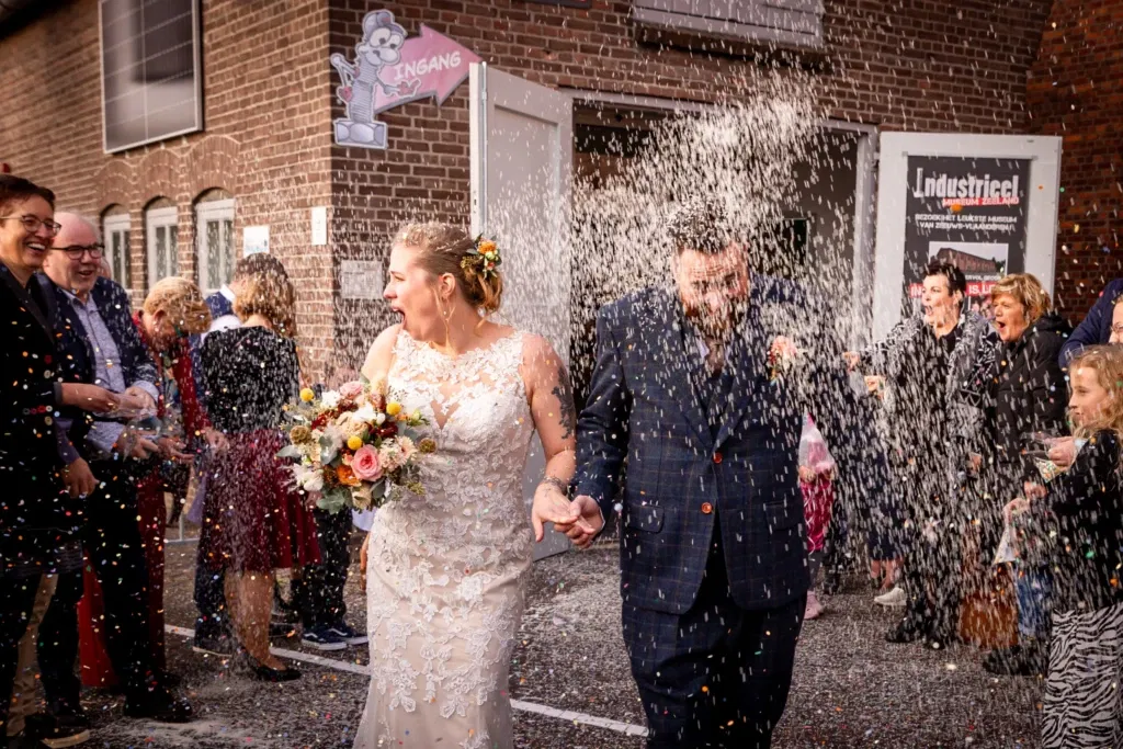 Een pasgetrouwd stel loopt hand in hand terwijl ze met vreugde door een regen van confetti gaan, omringd door lachende bruiloftsgasten. De bruid houdt een kleurrijk boeket vast en de bruidegom draagt een donkerblauw pak met een oranje boutonnière, passend bij de herfsttinten van het boeket.