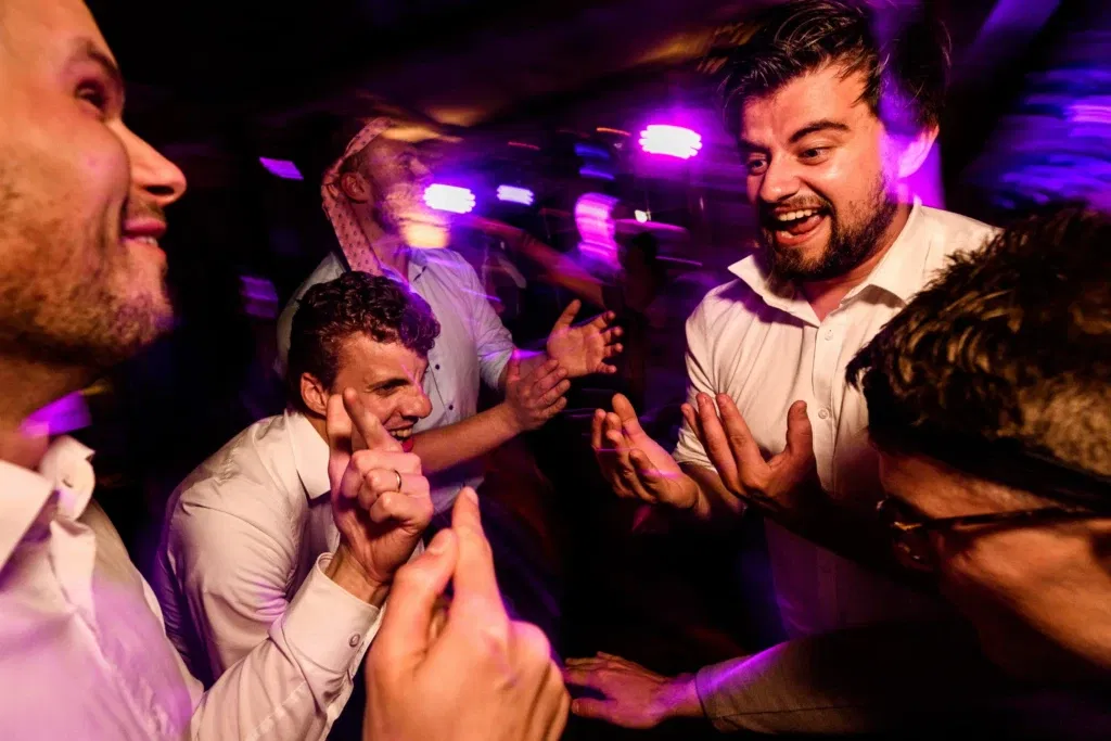 Een groep mannen in feeststemming, dansend en lachend onder gekleurd licht dat een gevoel van beweging en vrolijkheid uitstraalt op een dansvloer van bruidsfeest.