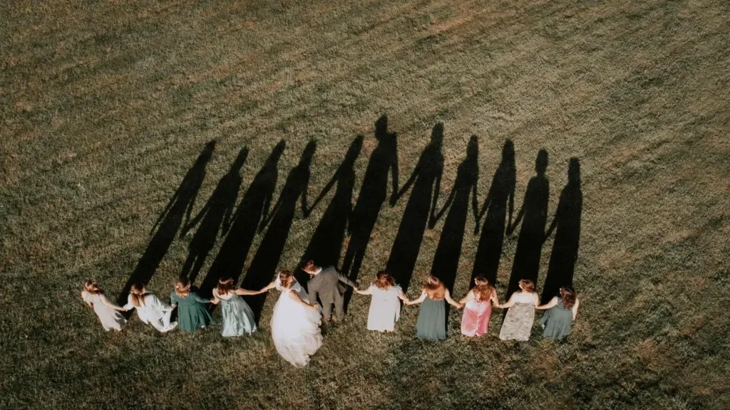 Een luchtfoto van een bruidspaar hand in hand met hun bruidsmeisjes, waarbij de lange schaduwen op het gras een lijn vormen