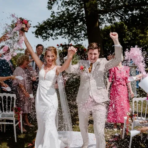 Een bruidspaar juicht enthousiast met opgeheven armen, terwijl ze onder een douche van roze bloemblaadjes en confetti doorlopen, omringd door vrolijke gasten op een zonnige dag.