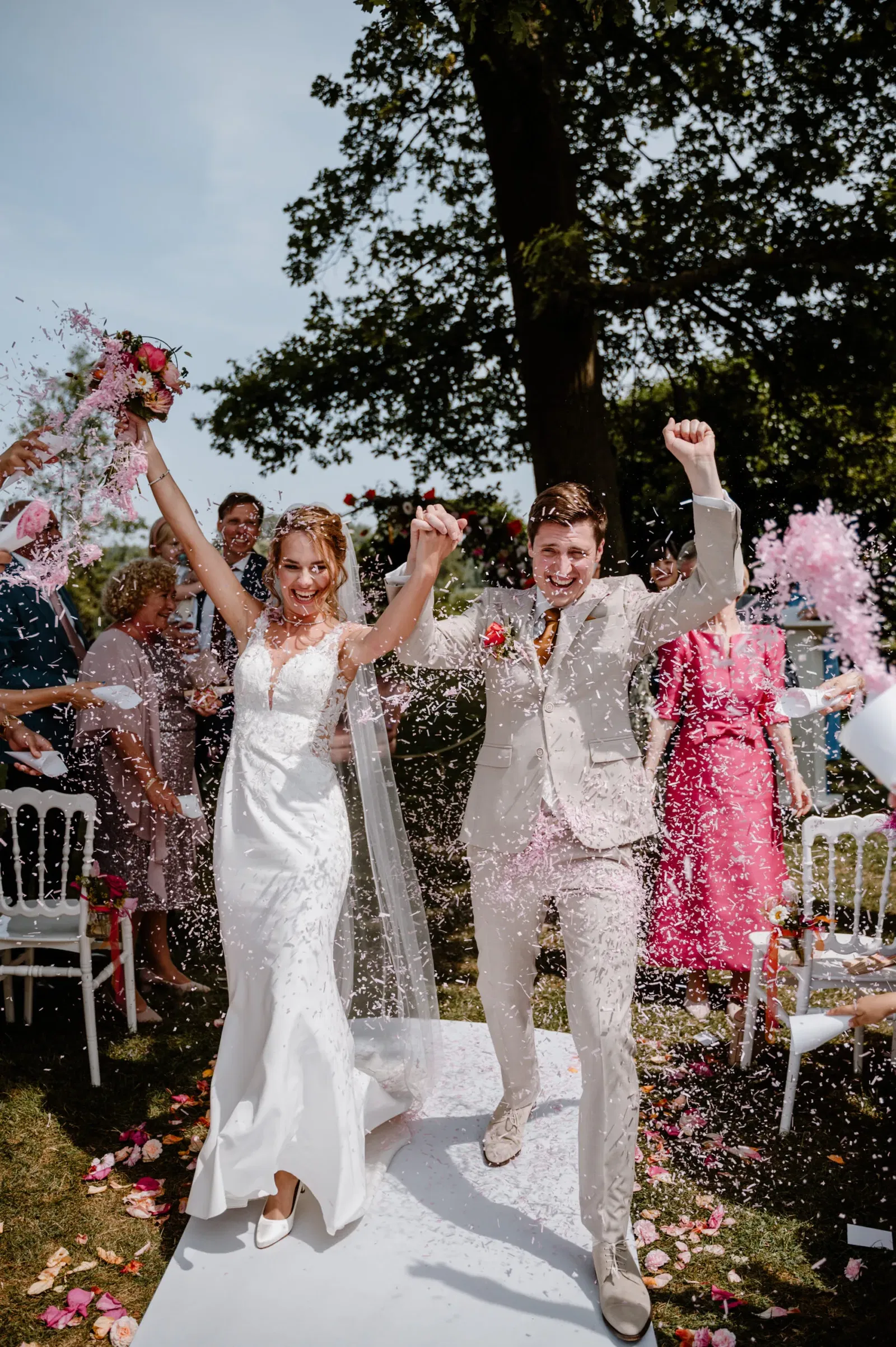 Een bruidspaar juicht enthousiast met opgeheven armen, terwijl ze onder een douche van roze bloemblaadjes en confetti doorlopen, omringd door vrolijke gasten op een zonnige dag.