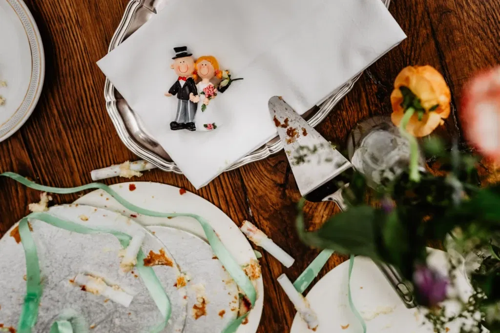 Een close-up van een bruidstaart topper met een miniatuur bruid en bruidegom naast de resten van de bruidstaart, met servetten, een taartschep en linten op een houten tafel.