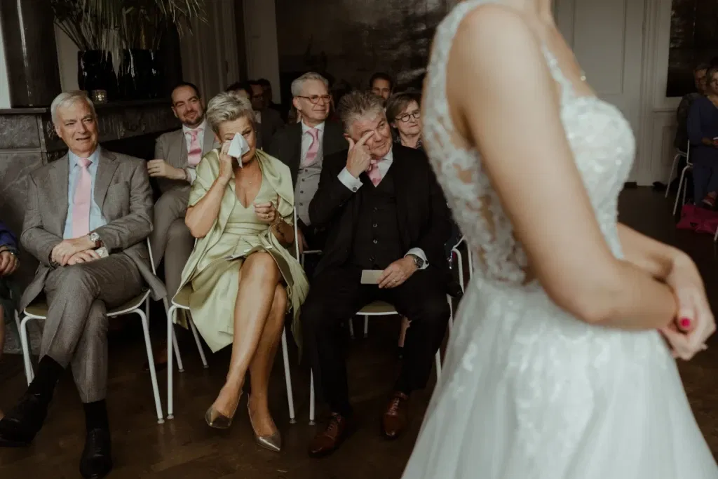 Bruiloftsgasten kijken emotioneel toe tijdens een ceremonie. Een vrouw in een lichtgroene jurk veegt tranen weg met een tissue. Op de voorgrond staat de bruid.