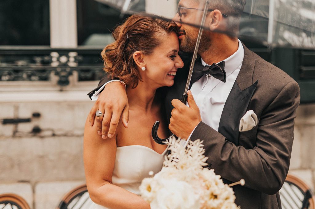 Bruidspaar onder paraplu. Bruidegom kust zijn bruid op haar voorhoofd.