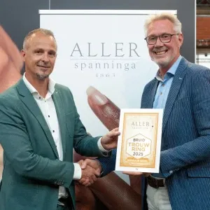 Twee mannen schudden elkaar de hand tijdens een prijsuitreiking. Eén van hen houdt een oorkonde vast met de tekst 'Aller Spanninga – Bruid Trouw Ring 2025 – Winnaar Goud'. Op de achtergrond is een banner van Aller Spanninga te zien