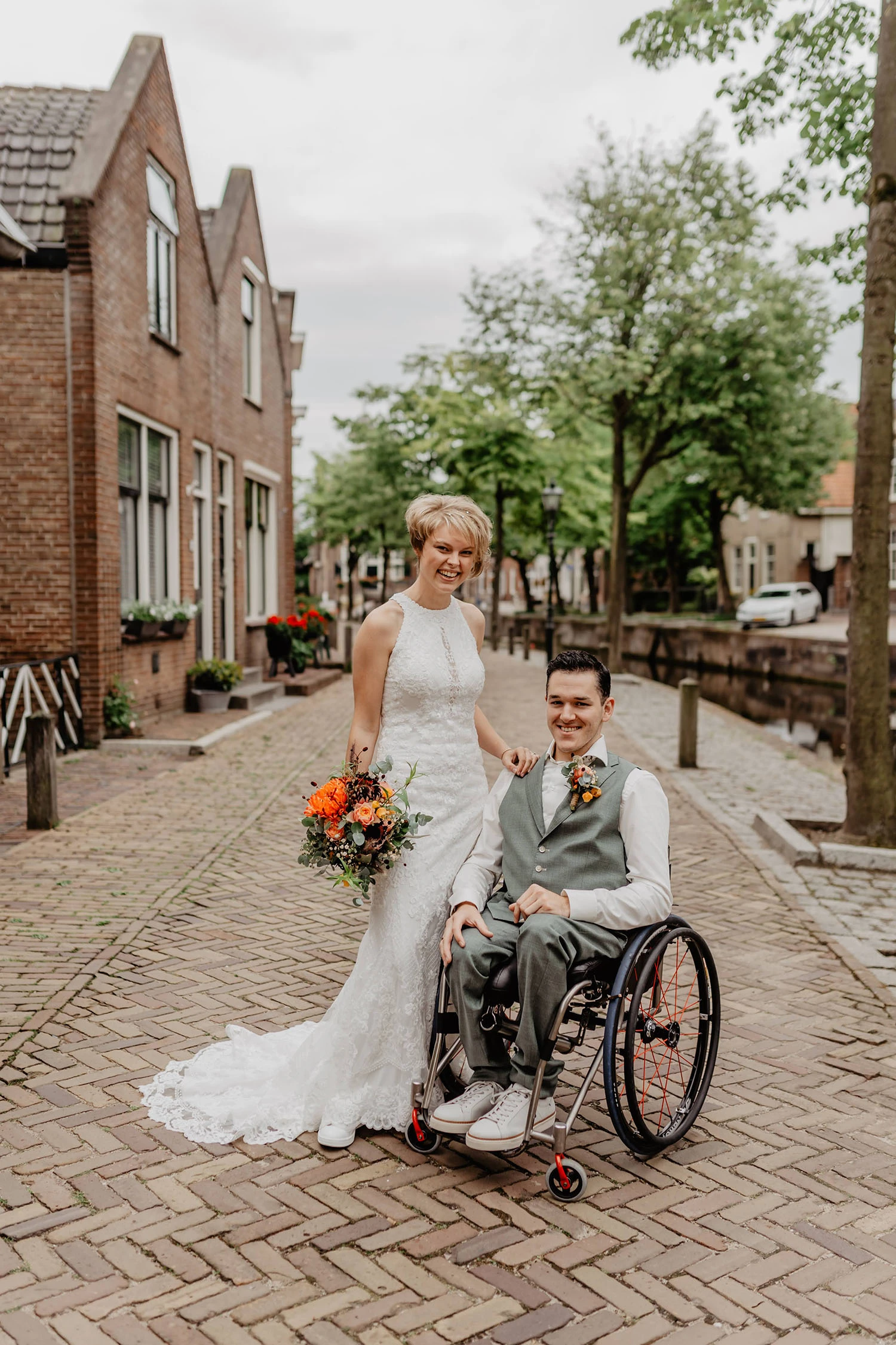 Bruidspaar poseert op een mooie straat. De bruid heeft een hoge gesloten hals witte jurk aan. De bruidegom zit in een rolstoel en heeft een mooi grijs gilet aan.