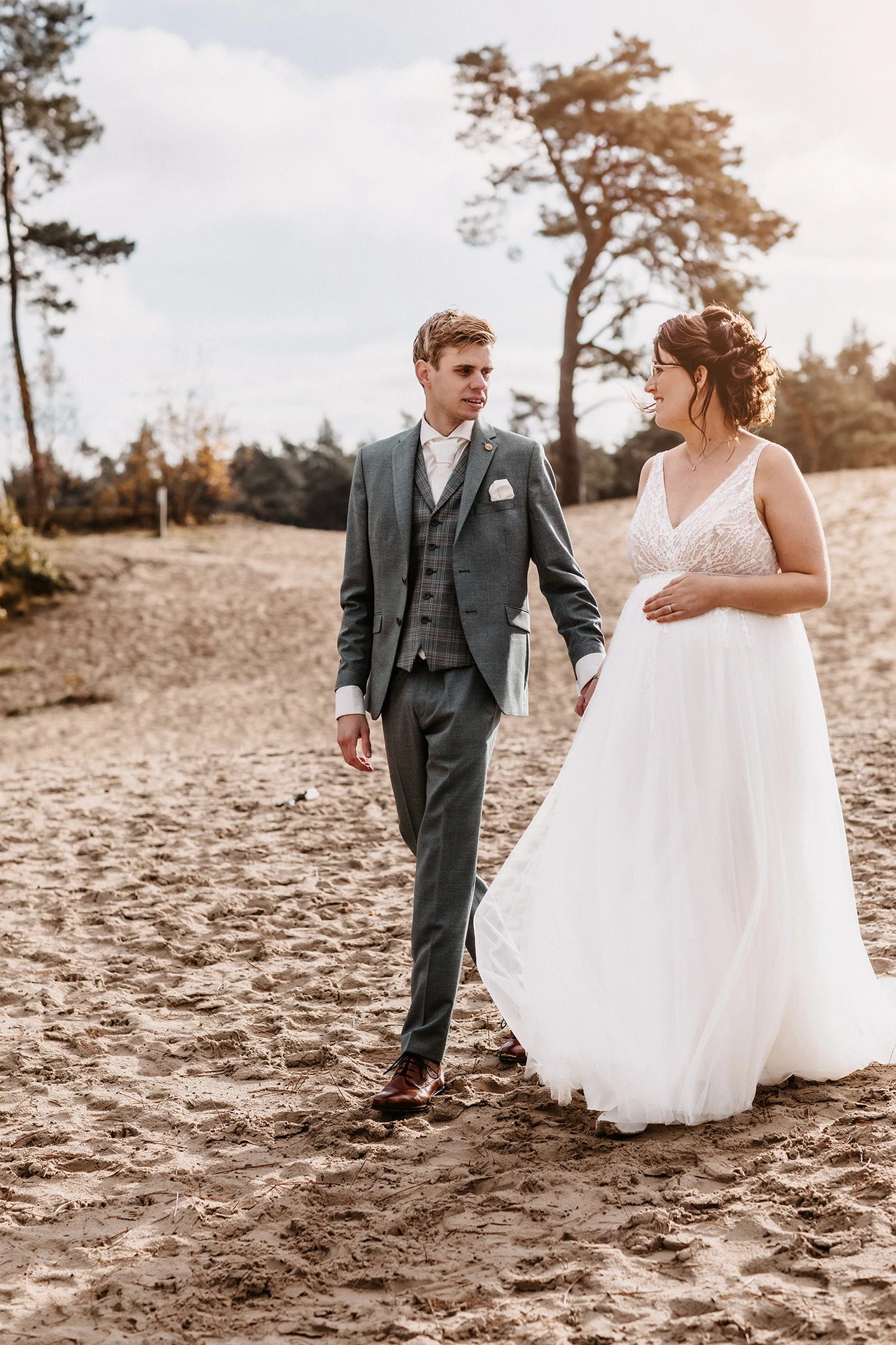 Bruidspaar loopt door de duinen. Bruid heeft een witte A-lijn jurk en houdt haar buik vast. Ze is zwanger. Bruidegom heeft een driedelig grijs pak aan. Ze lopen hand in hand.