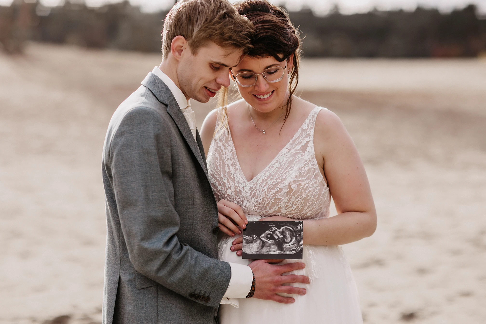 Bruidspaar met op de achtergrond duinen. Ze houden een echo foto vast van hun baby. De bruid is zwanger en draagt een mooie A-lijn trouwjurk. Bruidegom draagt een driedelig grijs pak