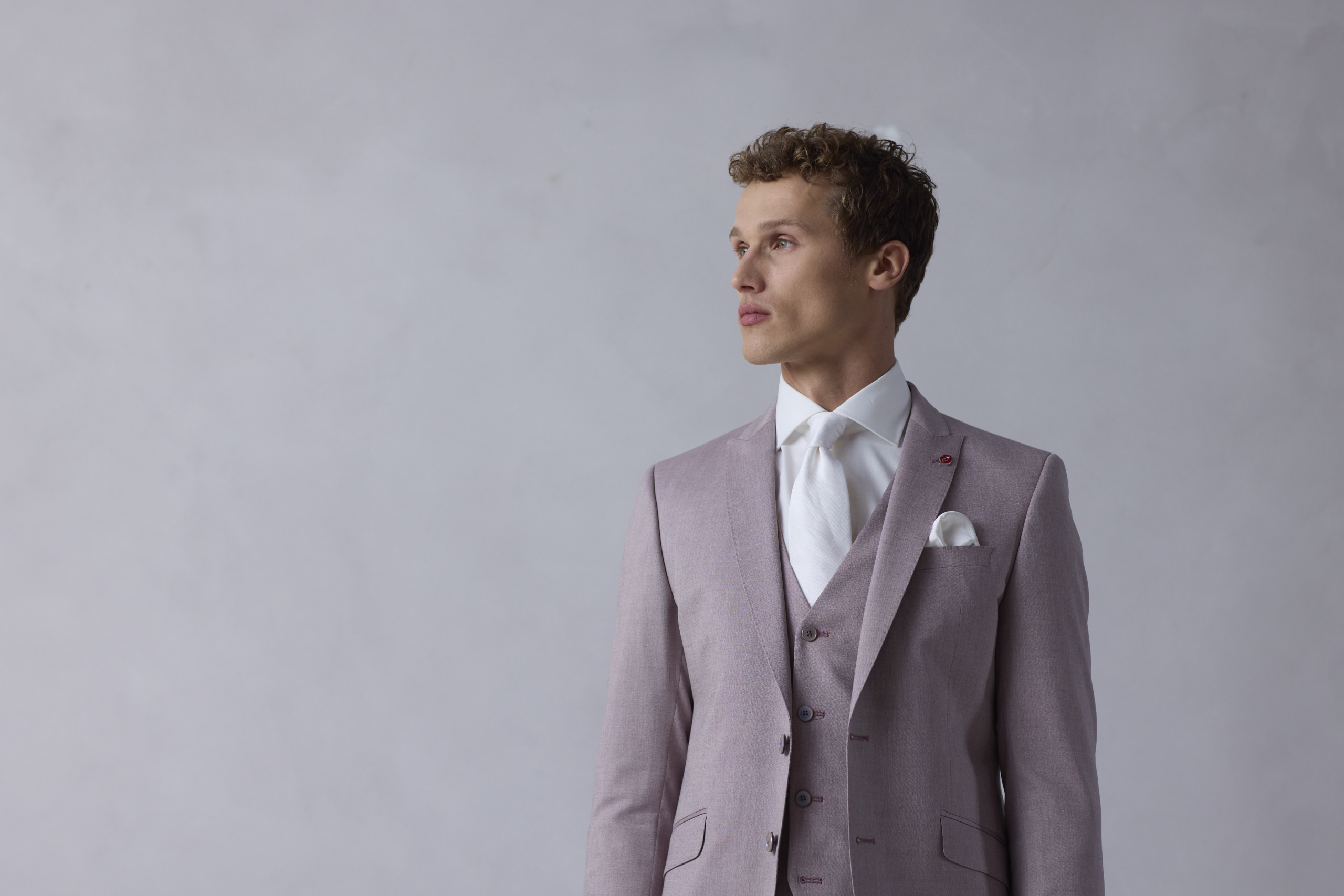 Jonge man met een driedelig trouwpak aan die een beetje rozig grijs is. Hij heeft een witte stropdas om en een wit pochet. De achtergrond is industrieel.