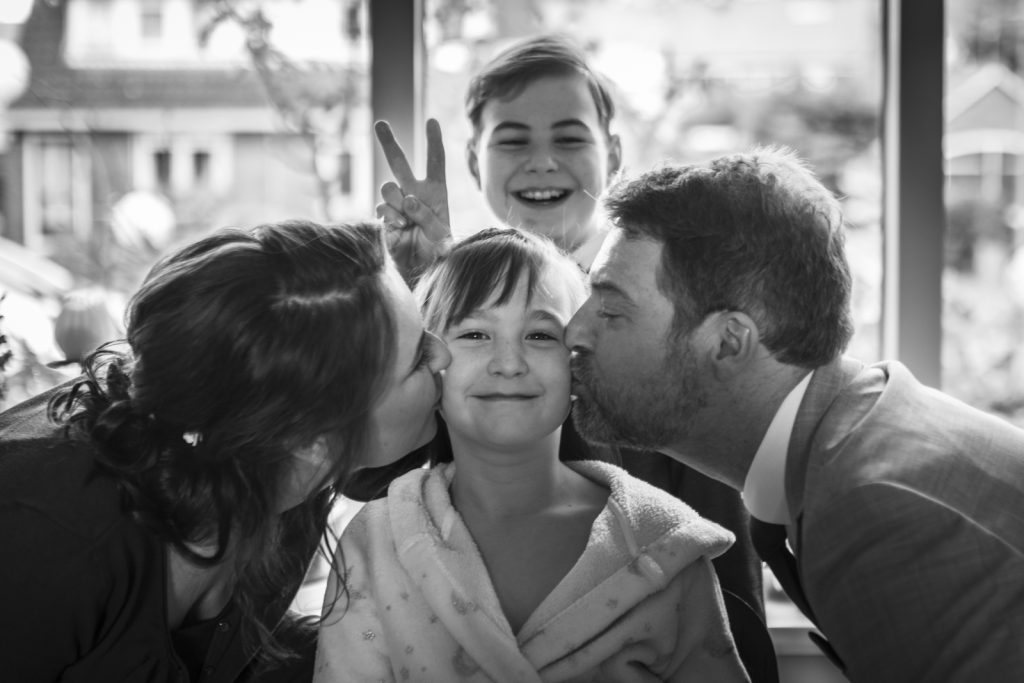 Een zwart-wit familieportret waarin een jong meisje glimlachend in het midden zit terwijl haar ouders haar liefdevol op de wangen kussen. Op de achtergrond staat een lachende jongen die vrolijk een peace-teken maakt met zijn vingers. De scène speelt zich af voor een raam met een wazige buitenomgeving.