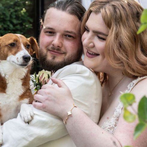 Bruidspaar dat poseert voor een huis. De bruid heeft een mooie witte jurk aan en staat achter de bruidegom. De bruidegom heeft een crèmekleurig pak aan en heeft hun bruin witte hondje in zijn handen.