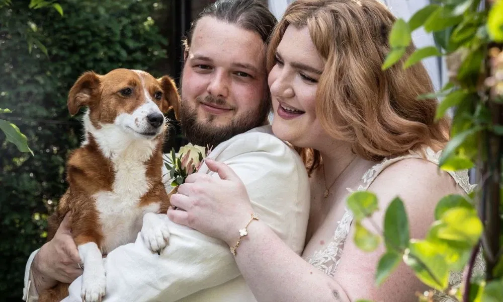 Bruidspaar dat poseert voor een huis. De bruid heeft een mooie witte jurk aan en staat achter de bruidegom. De bruidegom heeft een crèmekleurig pak aan en heeft hun bruin witte hondje in zijn handen.
