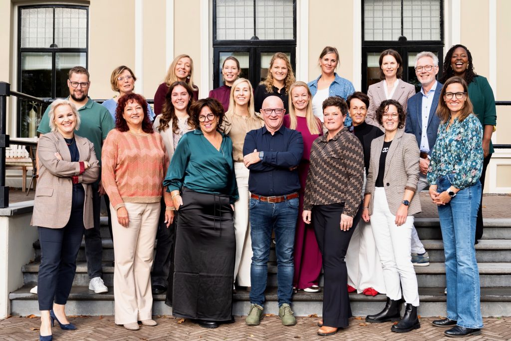 Een groep van twintig mensen poseert lachend voor een gebouw met een klassieke gevel en grote ramen. Ze staan op traptreden en dragen verschillende kledingstijlen, variërend van casual tot zakelijk. De sfeer is professioneel en vriendelijk.