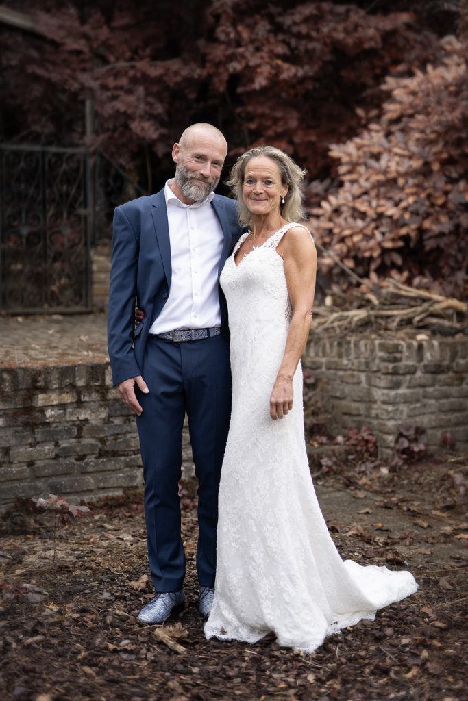 Bruidspaar poseert in een tuin. Bruid heeft een witte bruidsjurk met korte sleep aan. Bruidegom heeft een blauw pak met een wit overhemd aan.