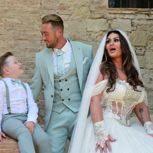 Bruid en bruidegom poseren voor een stenen muur in italië. Hun zoontje is ook aanwezig en de bruidegom kijkt hem lachend aan.