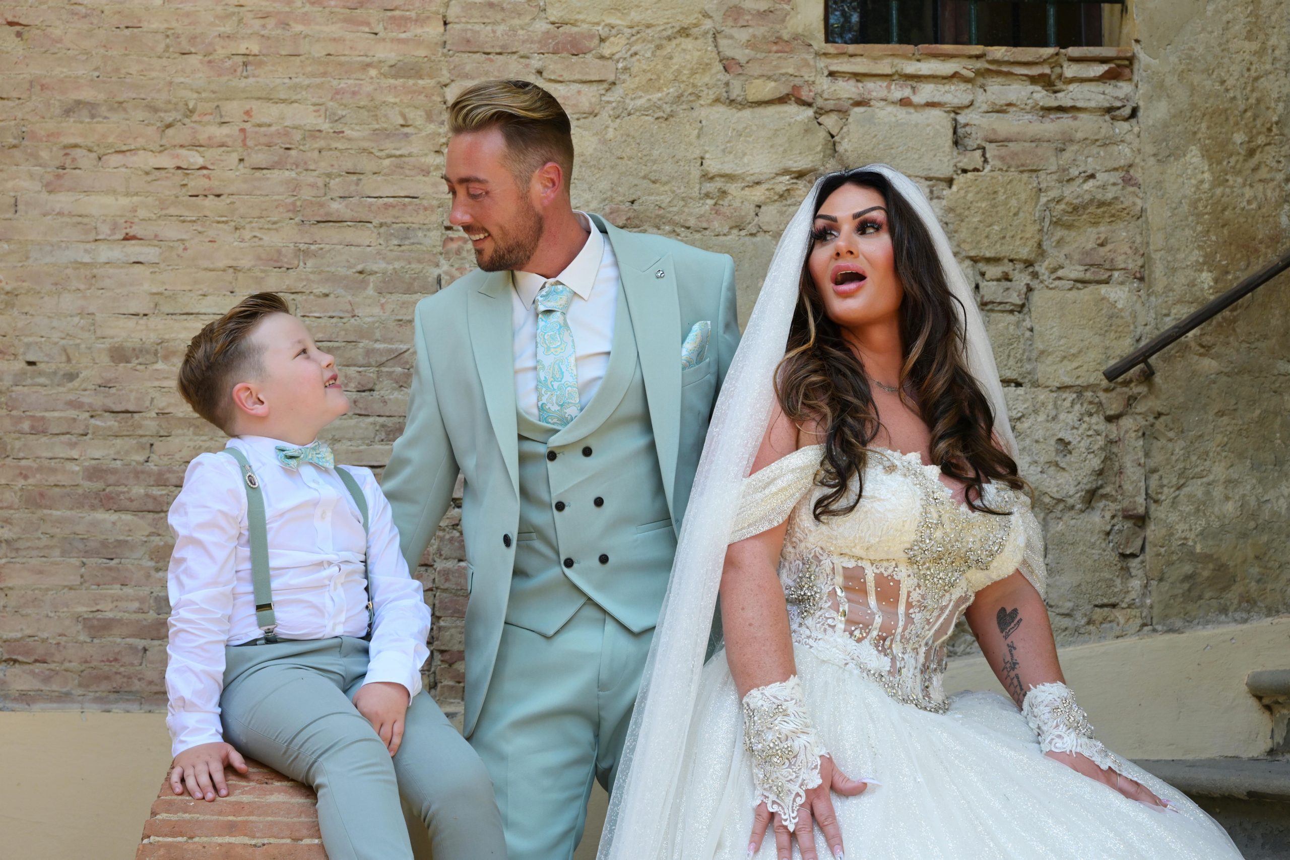 Bruid en bruidegom poseren voor een stenen muur in italië. Hun zoontje is ook aanwezig en de bruidegom kijkt hem lachend aan.