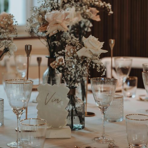 Een stijlvolle tafelopstelling met delicate glazen met gouden randjes, zachtgekleurde bloemen zoals rozen en gipskruid in glazen vazen, en een minimalistische tafeldecoratie in een warme, sfeervolle omgeving.
