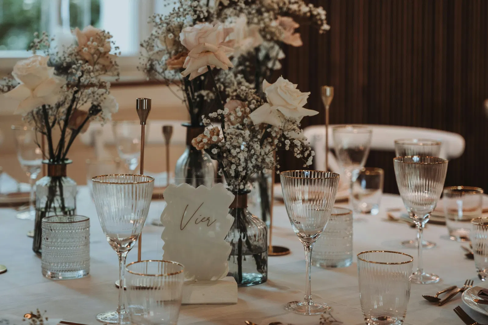 Een stijlvolle tafelopstelling met delicate glazen met gouden randjes, zachtgekleurde bloemen zoals rozen en gipskruid in glazen vazen, en een minimalistische tafeldecoratie in een warme, sfeervolle omgeving.