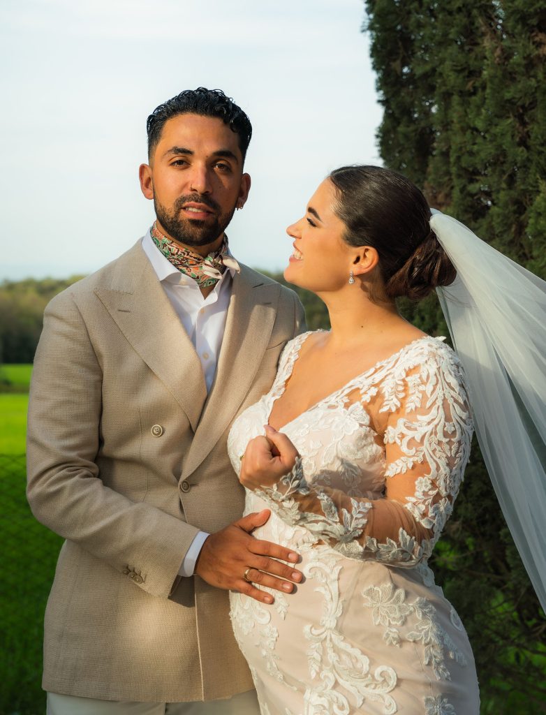 Bruidspaar poseert voor een grasvlakte. De bruid heeft een grijs pak aan. De bruid is zwanger en heeft een mooie witte strakke jurk aan. Bruidegom heeft hand op de buik en bruid lacht naar bruidegom.