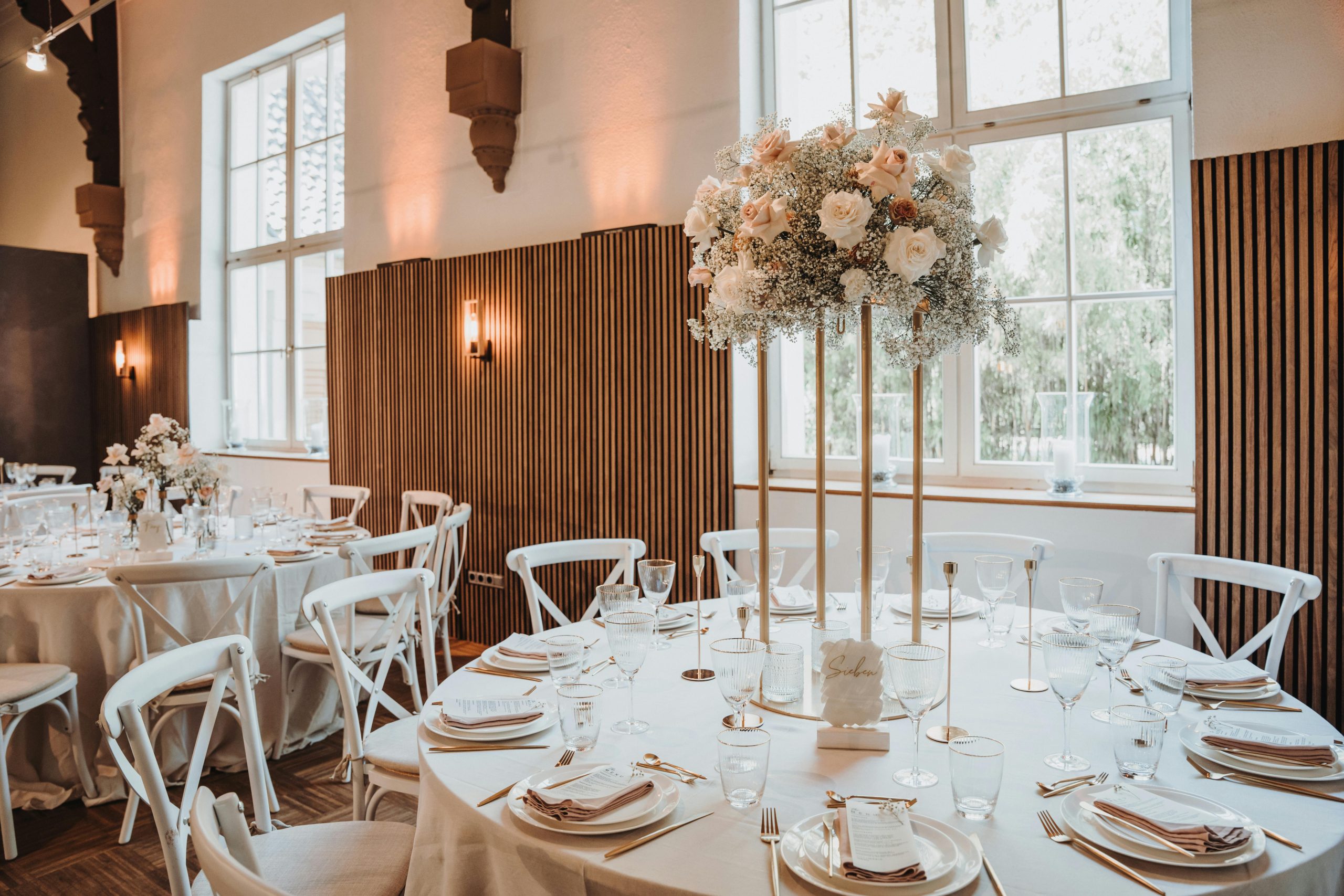 Elegante trouwlocatie met ronde tafels, gedecoreerd met witte tafelkleden, gouden bestek, kristallen glazen en een grote bloemenvaas met witte en pastelroze bloemen als centerpiece, omringd door witte stoelen in een lichte, stijlvolle zaal.