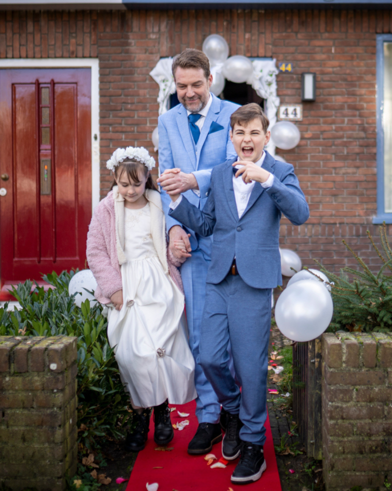 Een vader in een lichtblauw pak loopt trots met zijn twee kinderen over een rode loper voor hun huis, versierd met witte ballonnen. Het jonge meisje draagt een witte jurk met een roze jas en een bloemenkroon, terwijl de jongen in een blauw pak een speelse uitdrukking maakt. Het tafereel straalt vreugde en een feestelijke sfeer uit. Op de achtergrond is een rode voordeur en een bakstenen gevel te zien.