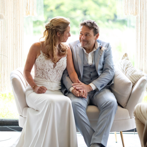 Een bruid en bruidegom zitten samen op een stijlvolle lichte bank, elkaar liefdevol aankijkend. De bruid draagt een elegante witte jurk met kant en de bruidegom een lichtblauw pak. Hun handen zijn in elkaar gevouwen, wat hun verbondenheid symboliseert. Op de achtergrond hangt een decoratief macramé gordijn, en buiten is een groene, rustige natuur zichtbaar.