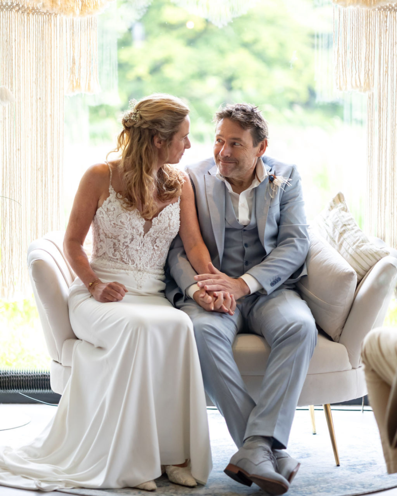 Een bruid en bruidegom zitten samen op een stijlvolle lichte bank, elkaar liefdevol aankijkend. De bruid draagt een elegante witte jurk met kant en de bruidegom een lichtblauw pak. Hun handen zijn in elkaar gevouwen, wat hun verbondenheid symboliseert. Op de achtergrond hangt een decoratief macramé gordijn, en buiten is een groene, rustige natuur zichtbaar.