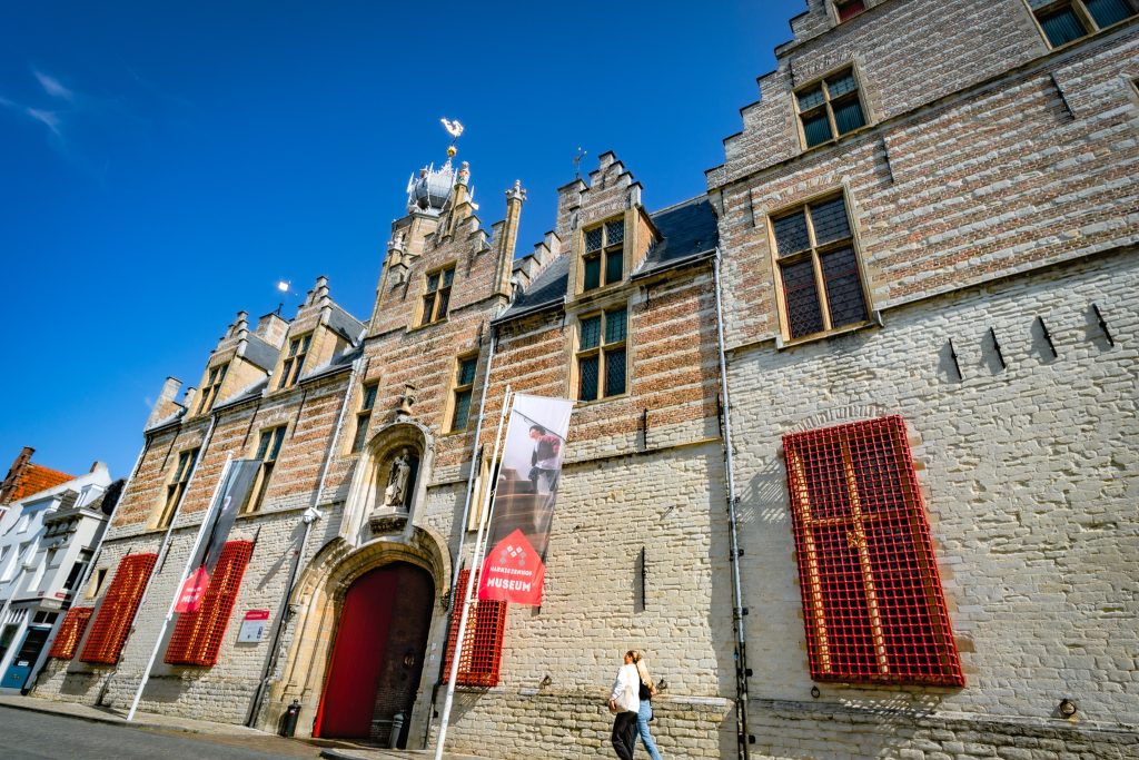 Foto van de historische voorgevel van het Markiezenhof in Bergen op Zoom, een laatgotisch stadspaleis met een indrukwekkende architectuur, rood-witte details en een grote rode toegangspoort. Twee voorbijgangers lopen langs het gebouw onder een stralend blauwe lucht, terwijl banners de ingang van het museum markeren.