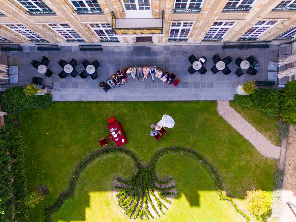 Luchtfoto van een elegante bruiloft in een Franse tuin, met een bruid in een witte jurk en een bruidegom aan een rood beklede tafel. De setting bevindt zich in de tuin van een historisch gebouw met een sierlijk aangelegd plantendesign, terwijl gasten op een terras toekijken.