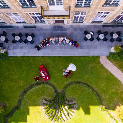 Luchtfoto van een elegante bruiloft in een Franse tuin, met een bruid in een witte jurk en een bruidegom aan een rood beklede tafel. De setting bevindt zich in de tuin van een historisch gebouw met een sierlijk aangelegd plantendesign, terwijl gasten op een terras toekijken.