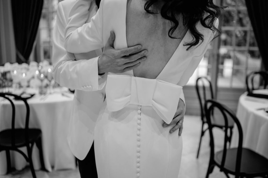 Zwart-wit close-up van een bruid en bruidegom die elkaar omhelzen tijdens hun huwelijksfeest. De bruid draagt een elegante jurk met een open rug, een grote strik en een rij knoopjes, terwijl de bruidegom in een wit pak haar liefdevol vasthoudt. Op de achtergrond zijn gedekte tafels en stoelen te zien in een sfeervolle trouwlocatie.