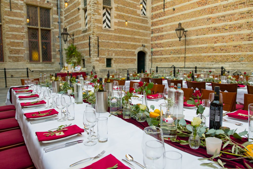 Een prachtig gedekte lange tafel op de Kleine Binnenplaats van een historisch gebouw, versierd met rode en witte accenten, groene takken, kaarsen en flessen wijn. De setting straalt een elegante en intieme sfeer uit, omgeven door middeleeuwse bakstenen muren en sfeerverlichting.